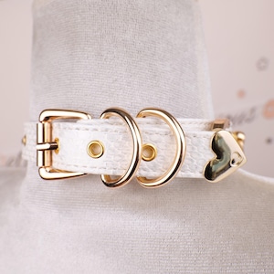 May include: White choker necklace with gold-colored hardware. The choker features a textured, snake-skin-like pattern, a gold heart charm, and a buckle closure. The metal hardware includes two D-rings and a buckle.