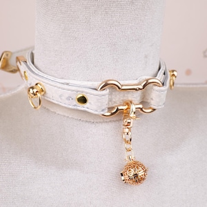 May include: White faux leather choker necklace with gold-colored hardware. The choker features a textured pattern, a gold-colored chain, and a decorative gold-colored filigree ball pendant. The necklace is fastened with a buckle.