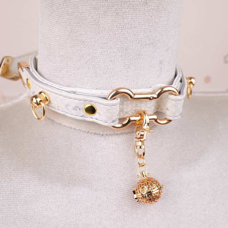 May include: White faux leather choker necklace with gold-colored hardware. The choker features a textured pattern, a gold-colored chain, and a decorative gold-colored filigree ball pendant. The necklace is fastened with a buckle.