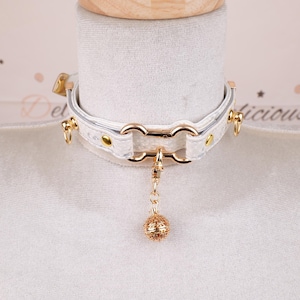 May include: White choker necklace with gold-colored hardware and a dangling gold ball charm. The choker features a textured pattern and is fastened with a buckle. The necklace is displayed on a white mannequin.