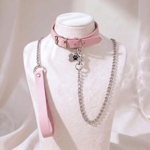 Op de afbeelding: Een lichtroze halsband met een zilverkleurige gesp en een bijpassende riem met een zilveren metalen ketting. Een kleine zilveren botvormige hanger met een zwarte pootafdruk hangt aan de D-ring van de halsband en verbindt met de ketting.