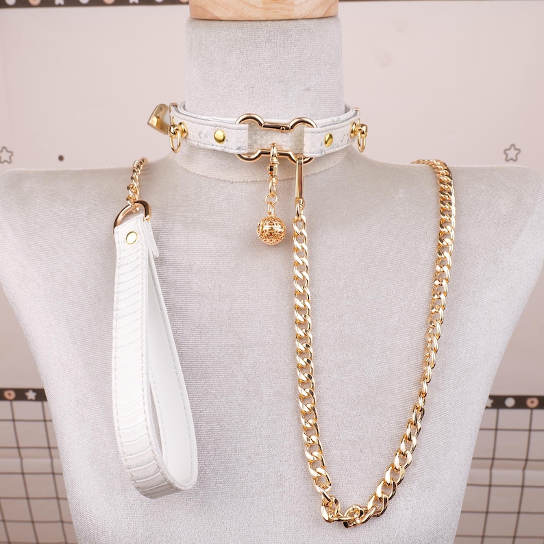 May include: White leather collar with gold-colored hardware, including a chain leash and a decorative charm. The collar features a lock and is displayed on a mannequin.