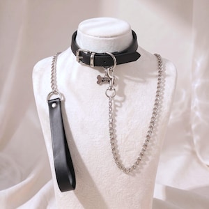 May include: A black leather collar featuring a silver-toned metal buckle and a D-ring. A silver-toned metal chain leash with a black leather handle is attached to the collar. A small silver-toned bone-shaped charm hangs from the D-ring.