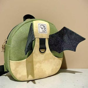 Puede incluir: Una mochila verde y beige con alas de dragón negras. La mochila tiene un parche de dinosaurio de dibujos animados y un pequeño bolsillo lateral. Las alas de color gris oscuro añaden un toque lúdico al diseño.