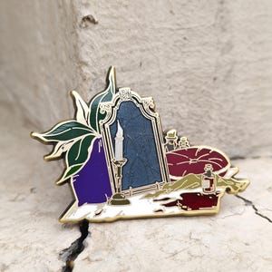 May include: A gold-toned metal enamel pin featuring a central arched mirror with a cracked surface, a lit candle in a holder, a dark purple plant, a maroon circular cushion, and small glass potion bottles, all outlined in polished gold metal.