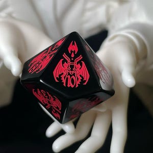 May include: A black, multi-sided die with red, stylized illustrations and the number 10. The die is held in gloved hands against a blurred background. The illustrations appear to be of a fantasy or gothic theme.