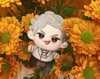 May include: A plush doll with gray hair, red eyes, and a white shirt with a black vest, surrounded by orange and yellow flowers. The doll has a playful expression with its tongue sticking out.