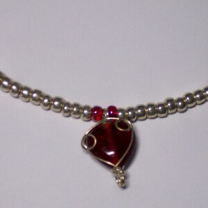 May include: A silver beaded necklace with red detailing. The necklace has a heart-shaped pendant and two circular charms with red centres. Small red beads are spaced along the chain.