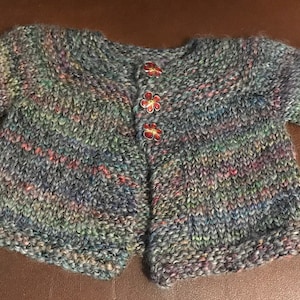 May include: A hand-knitted baby cardigan in a mix of colors including blue, green, and pink. The sweater has three flower-shaped buttons and rolled cuffs on the sleeves. The yarn appears to be a soft, textured blend.