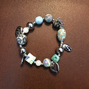 Puede incluir: Una pulsera de cuentas con una variedad de cuentas y abalorios coloridos. La pulsera presenta cuentas de metal plateado, cuentas blancas y cuentas estampadas en colores azul, verde y crema. Los abalorios incluyen un corazón, una huella y un pequeño colgante.