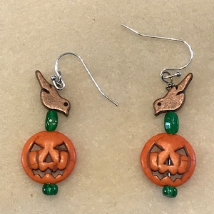 Op de afbeelding: Een paar Halloween-oorbellen. Elke oorbel heeft een oranje pompoen kraal met een uitgesneden jack-o'-lantern gezicht, een groene kraal en een koperkleurige vogelbedel. De oorbellen hebben zilverkleurige oorhaken.