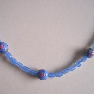 Pode incluir: Um colar de contas azul claro com três contas redondas maiores. Cada conta redonda tem um design em espiral rosa e branco. O colar é feito de pequenas contas ovais.