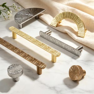 May include: A collection of decorative cabinet hardware in various metallic finishes. The selection includes handles and knobs with textured, linear and radial designs. The hardware is in silver, gold, and bronze tones, displayed on a marble surface.