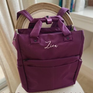 May include: Plum-colored backpack with a top handle and adjustable shoulder straps. The bag features a front pocket and a zippered main compartment. The name "Ziva" is embroidered in light pink script on the front. The bag is sitting on a chair.