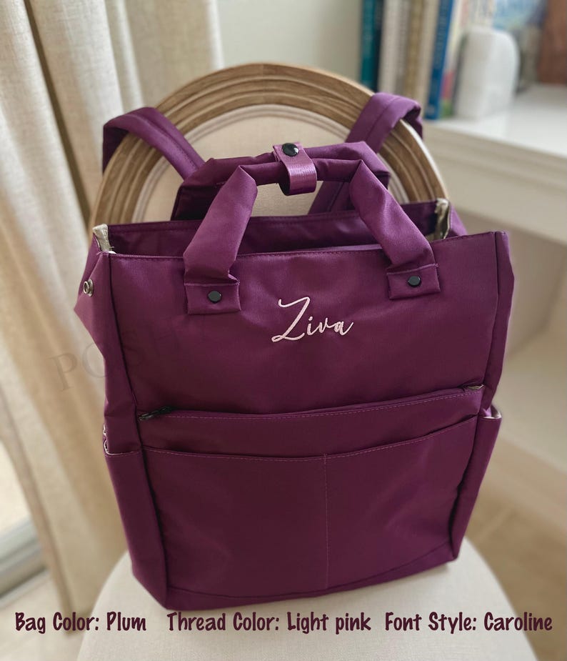 May include: Plum-colored backpack with a top handle and adjustable shoulder straps. The bag features a front pocket and a zippered main compartment. The name "Ziva" is embroidered in light pink script on the front. The bag is sitting on a chair.