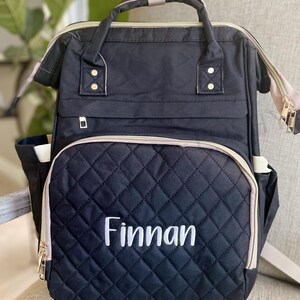 May include: Black quilted backpack with the name "Finnan" embroidered in white. The bag has a top handle, adjustable straps, and multiple pockets. Gold zippers and accents add detail to the design.