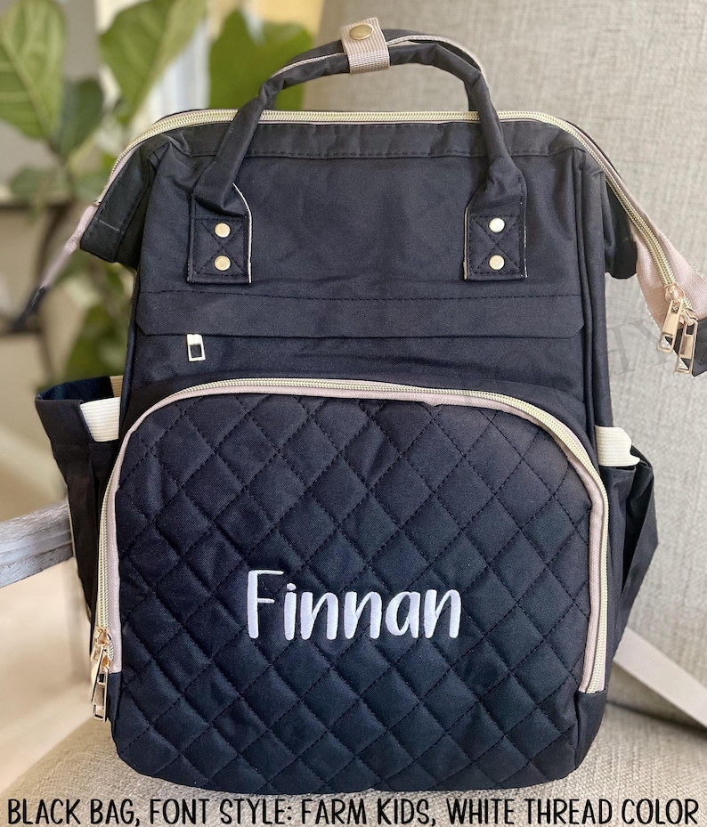 May include: Black quilted backpack with the name "Finnan" embroidered in white. The bag has a top handle, adjustable straps, and multiple pockets. Gold zippers and accents add detail to the design.