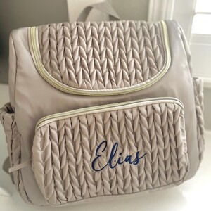 May include: A beige backpack with a quilted design and gold-colored zippers. The front pocket features the name "Elias" embroidered in blue script. The bag has a top handle and side pockets, ideal for carrying essentials.