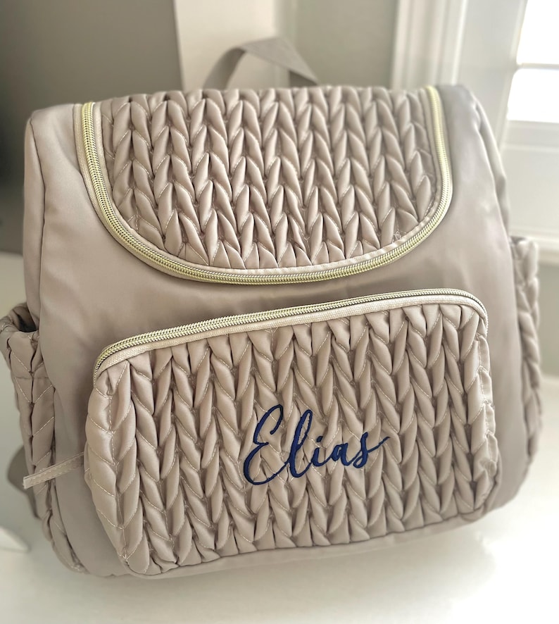May include: A beige backpack with a quilted design and gold-colored zippers. The front pocket features the name "Elias" embroidered in blue script. The bag has a top handle and side pockets, ideal for carrying essentials.