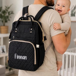May include: Black diaper bag with gold-colored zippers and trim. The front pocket has a quilted pattern and the name "Finnan" embroidered in white. The bag is being worn as a backpack.