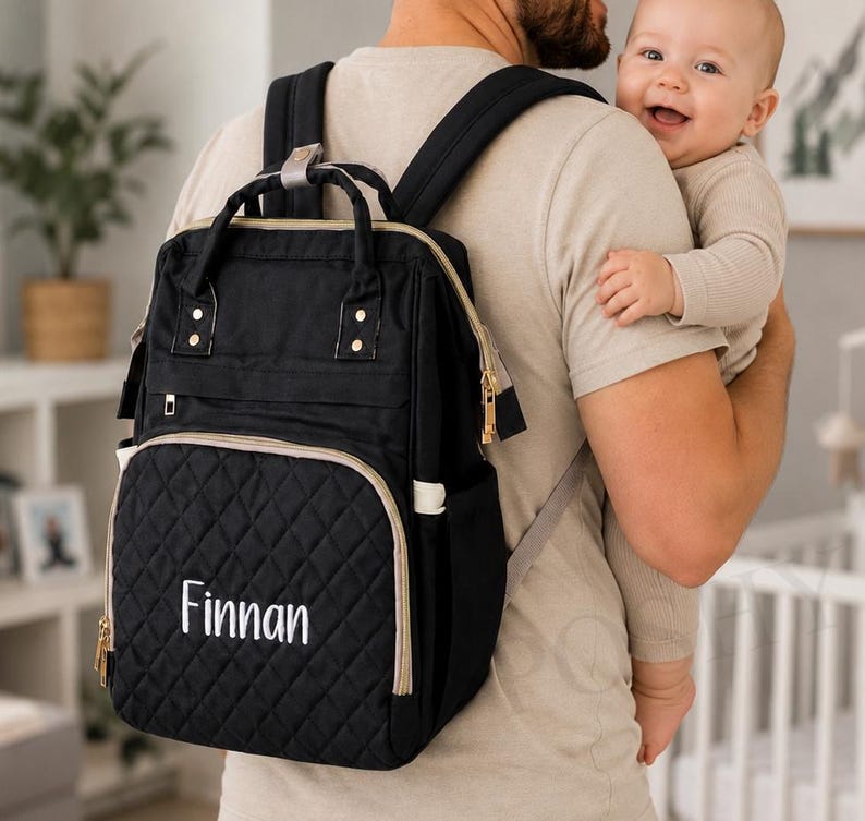 May include: Black diaper bag with gold-colored zippers and trim. The front pocket has a quilted pattern and the name "Finnan" embroidered in white. The bag is being worn as a backpack.