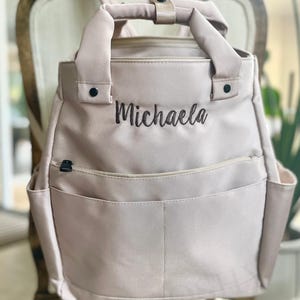 May include: A light pink backpack with the name "Michaela" embroidered in black. The bag has a top handle, adjustable straps, and multiple pockets, including side pockets and a front pocket. The backpack is sitting on a wooden chair.