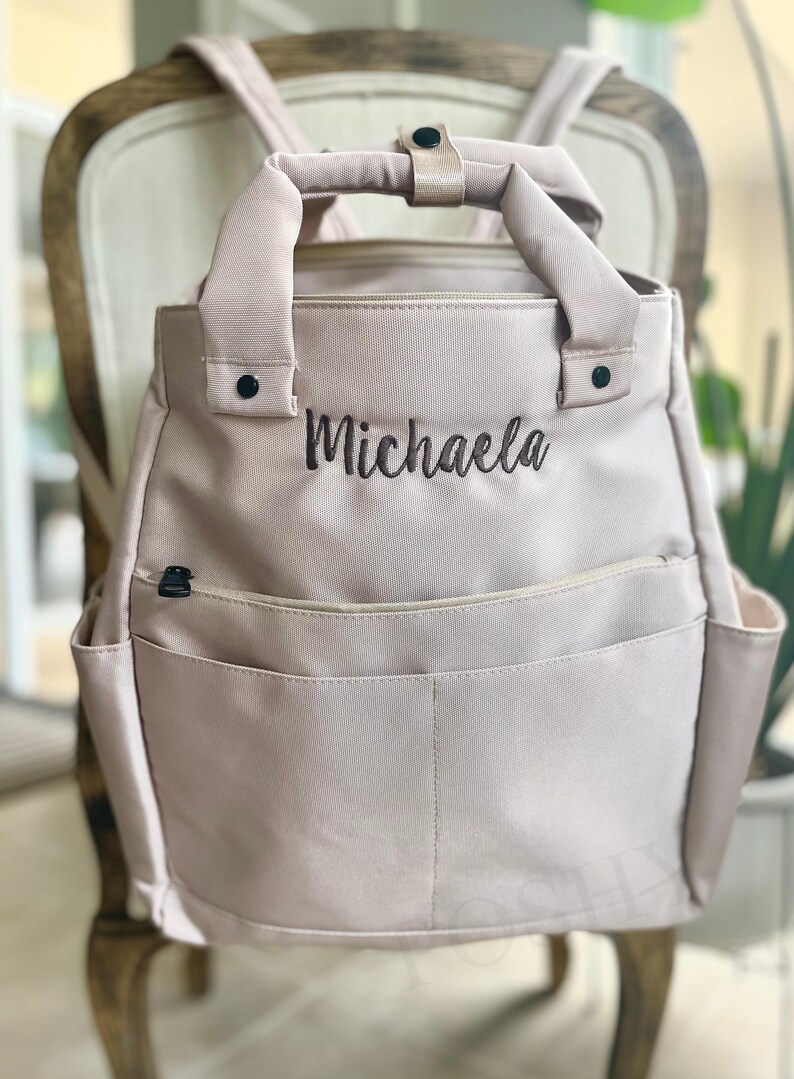 May include: A light pink backpack with the name "Michaela" embroidered in black. The bag has a top handle, adjustable straps, and multiple pockets, including side pockets and a front pocket. The backpack is sitting on a wooden chair.