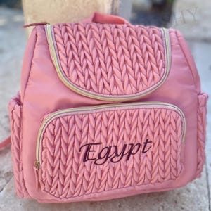 May include: Pink backpack with a quilted design on the front pocket and flap. The word "Egypt" is embroidered in black script on the front pocket. The backpack has gold zippers and a top handle.