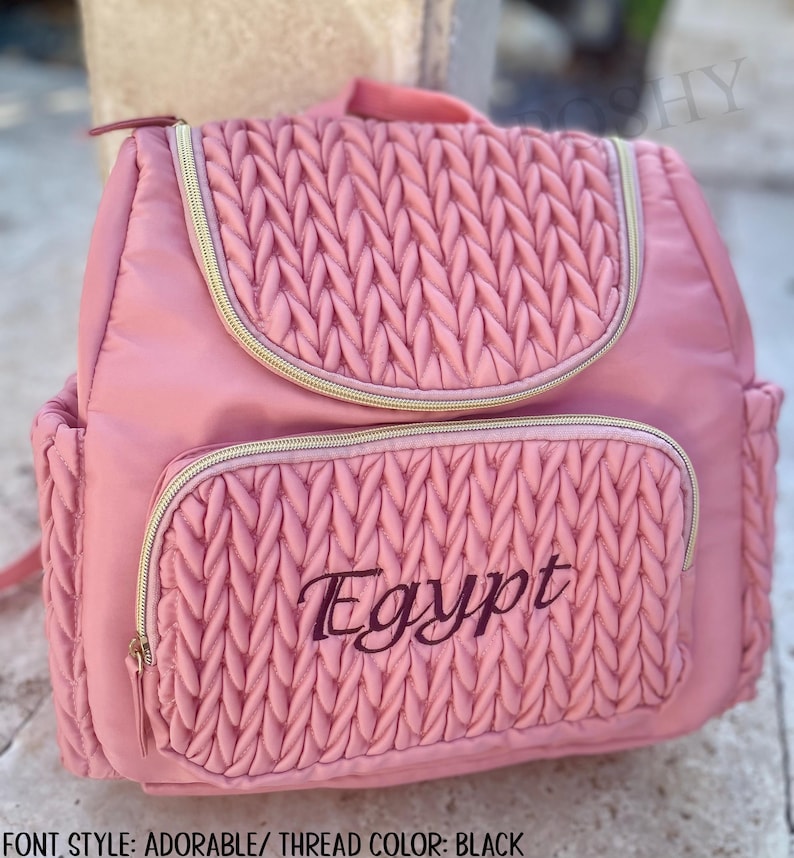 May include: Pink backpack with a quilted design on the front pocket and flap. The word "Egypt" is embroidered in black script on the front pocket. The backpack has gold zippers and a top handle.