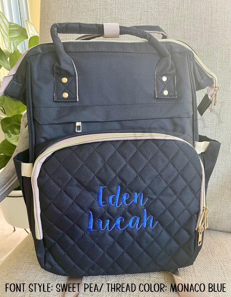 May include: Black quilted diaper bag with blue embroidered text reading "Eden Lucah." The bag has gold zippers, a light gray trim, and a top handle. The bag has a front pocket and side pockets. The text at the bottom reads "FONT STYLE: SWEET PEA/ THREAD COLOR: MONACO BLUE."