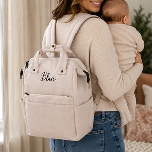 May include: A beige backpack with the name "Blair" in cursive script. The backpack has a top handle, adjustable straps, and multiple pockets. The person in the image is wearing blue jeans and holding a baby.