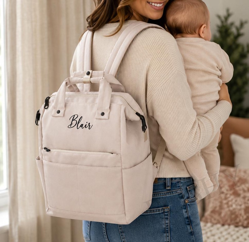 May include: A beige backpack with the name "Blair" in cursive script. The backpack has a top handle, adjustable straps, and multiple pockets. The person in the image is wearing blue jeans and holding a baby.