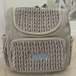 May include: A gray backpack with a quilted front pocket and flap, featuring a gold zipper and a top handle. The name "Laidyn" is embroidered in blue script on the front pocket. The bag has side pockets and a padded back.