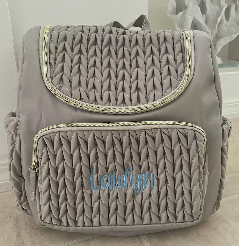May include: A gray backpack with a quilted front pocket and flap, featuring a gold zipper and a top handle. The name "Laidyn" is embroidered in blue script on the front pocket. The bag has side pockets and a padded back.