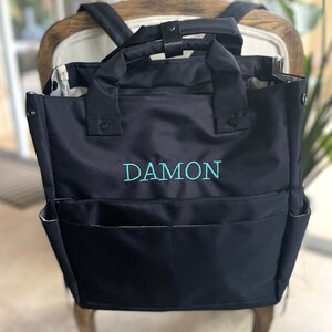 May include: Black backpack-style tote bag with teal embroidered text "DAMON". The bag has two exterior pockets and two handles. The interior lining is patterned. The bag is sitting on a chair.