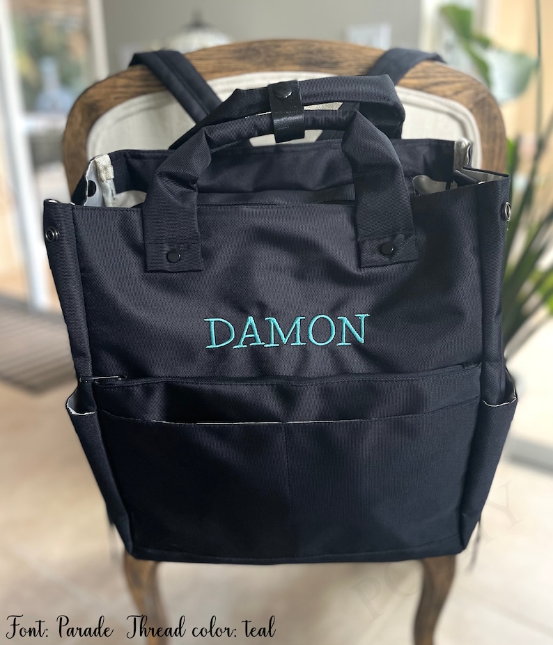 May include: Black backpack-style tote bag with teal embroidered text "DAMON". The bag has two exterior pockets and two handles. The interior lining is patterned. The bag is sitting on a chair.