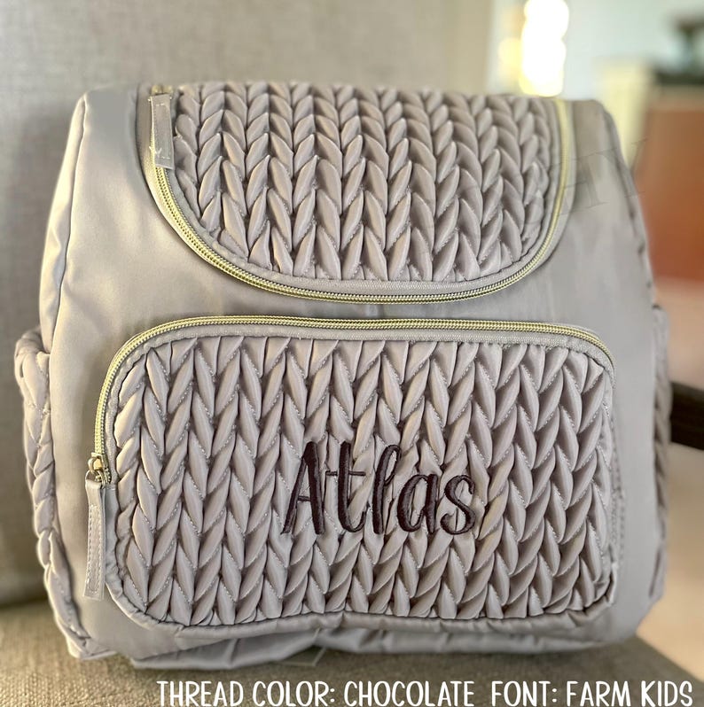 May include: A gray backpack with a quilted design on the front pocket and flap. The name "Atlas" is embroidered in chocolate-colored thread. The backpack has gold-colored zippers and a top handle.