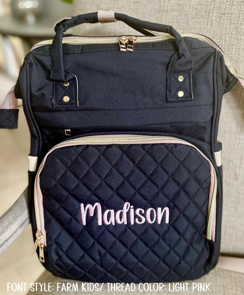 May include: A navy blue quilted backpack with a light pink trim and zipper. The front pocket features the name "Madison" embroidered in light pink. The bag has a top handle and adjustable straps, ideal for carrying baby essentials.