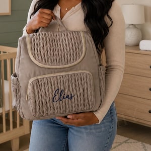 May include: A taupe-colored diaper bag with a quilted design and gold trim. The bag features a front pocket with the name "Elias" embroidered in navy blue. It has a top handle and side pockets.