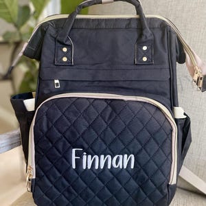 以下が含まれることがあります： 白で「Finnan」と刺繍された黒のキルティングバックパック。バッグには上部のハンドル、調節可能なストラップ、複数のポケットが付いています。ゴールドのジッパーとアクセントがデザインにディテールを追加します。ベビー用品の持ち運び用に設計されています。