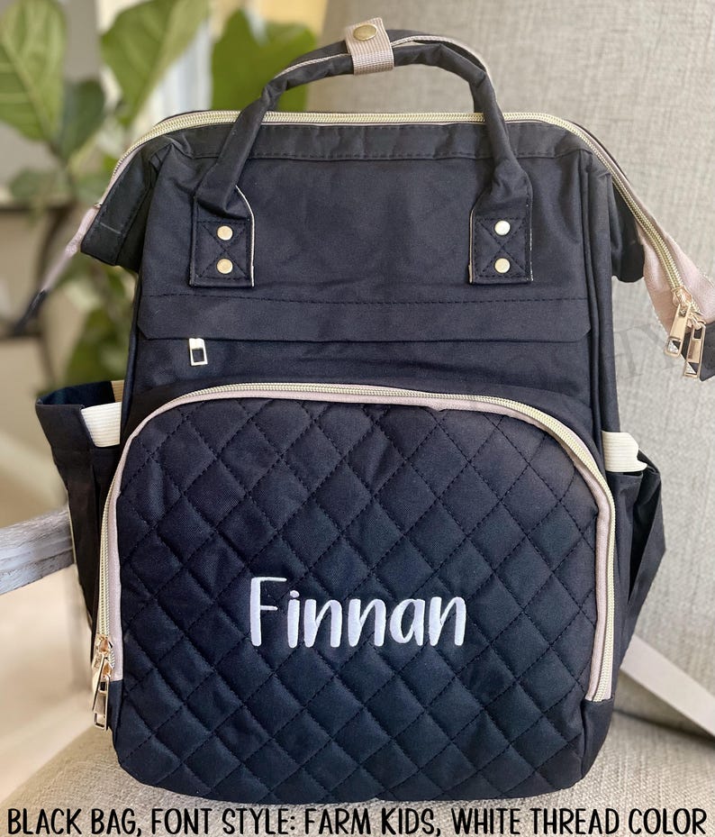 以下が含まれることがあります： 白で「Finnan」と刺繍された黒のキルティングバックパック。バッグには上部のハンドル、調節可能なストラップ、複数のポケットが付いています。ゴールドのジッパーとアクセントがデザインにディテールを追加します。ベビー用品の持ち運び用に設計されています。