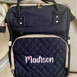 以下が含まれることがあります： 前面のポケットにライトピンクで「Madison」と刺繍された、ネイビーブルーのキルティングバックパック。ゴールドのジッパーとアクセント、トップハンドル、調節可能なストラップが付いています。ベビー用品の持ち運びに適しています。