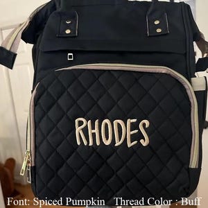 以下が含まれることがあります： 黒色のキルティングおむつバッグ。名前「RHODES」がバフ色の糸で刺繍されています。バッグには、ゴールドのジッパーとタン色のトリムが付いたフロントポケットがあります。上部にハンドルと調節可能なショルダーストラップが付いています。