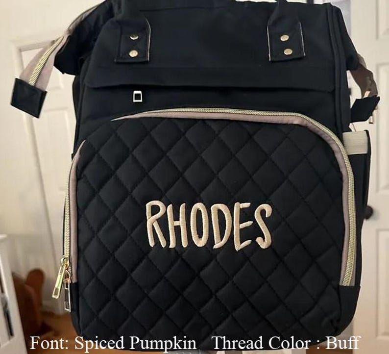 以下が含まれることがあります： 黒色のキルティングおむつバッグ。名前「RHODES」がバフ色の糸で刺繍されています。バッグには、ゴールドのジッパーとタン色のトリムが付いたフロントポケットがあります。上部にハンドルと調節可能なショルダーストラップが付いています。
