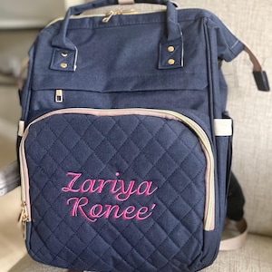 May include: Navy blue diaper bag with a quilted front panel. The bag features a pink zipper and trim, and the name "Zariya Ronee" is embroidered in bright pink. It has multiple compartments and a top handle.