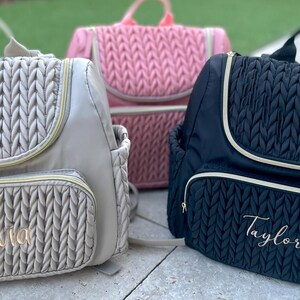 May include: Three diaper backpacks in beige, pink, and black, each with a quilted design. The beige and black bags have a gold zipper and trim, with the names "Olivia" and "Taylor" embroidered on the front pocket.