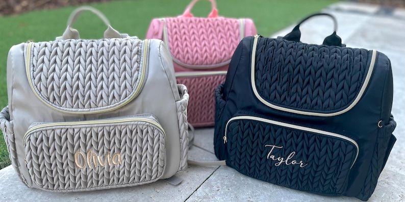 May include: Three diaper backpacks in beige, pink, and black, each with a quilted design. The beige and black bags have a gold zipper and trim, with the names "Olivia" and "Taylor" embroidered on the front pocket.