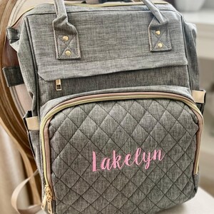 May include: Gray quilted diaper bag with the name "Lakelyn" embroidered in pink script. The bag has multiple compartments, gold zippers, and a top handle. The bag is sitting on a chair.