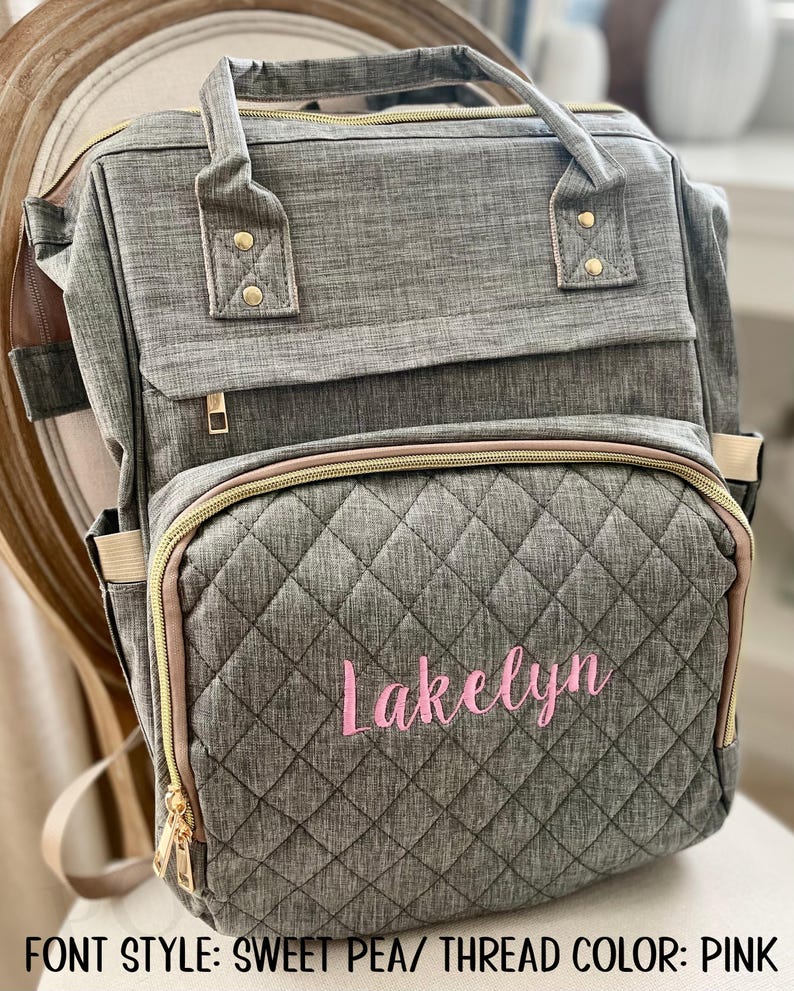 May include: Gray quilted diaper bag with the name "Lakelyn" embroidered in pink script. The bag has multiple compartments, gold zippers, and a top handle. The bag is sitting on a chair.