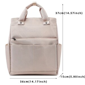 May include: A beige backpack with a top handle and two side pockets. The bag is 14.17 inches wide, 14.57 inches tall, and 5.90 inches deep. It has a front zippered pocket and two snap closures on the handle.